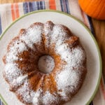 A pumpkin bundt cake dusted with powdered sugar on a plate next to two pumpkins.