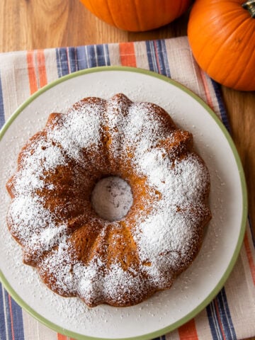 A pumpkin bundt cake dusted with powdered sugar on a plate next to two pumpkins.