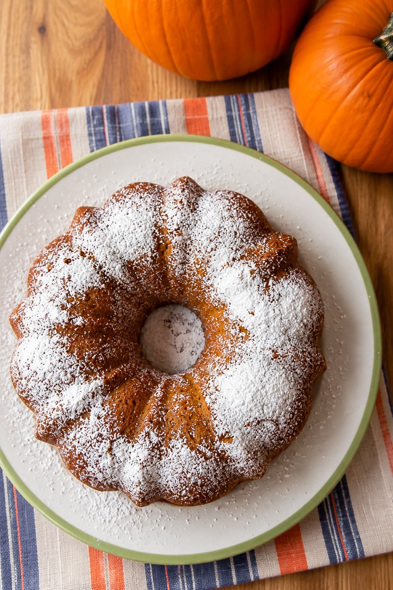A pumpkin bundt cake dusted with powdered sugar on a plate next to two pumpkins.