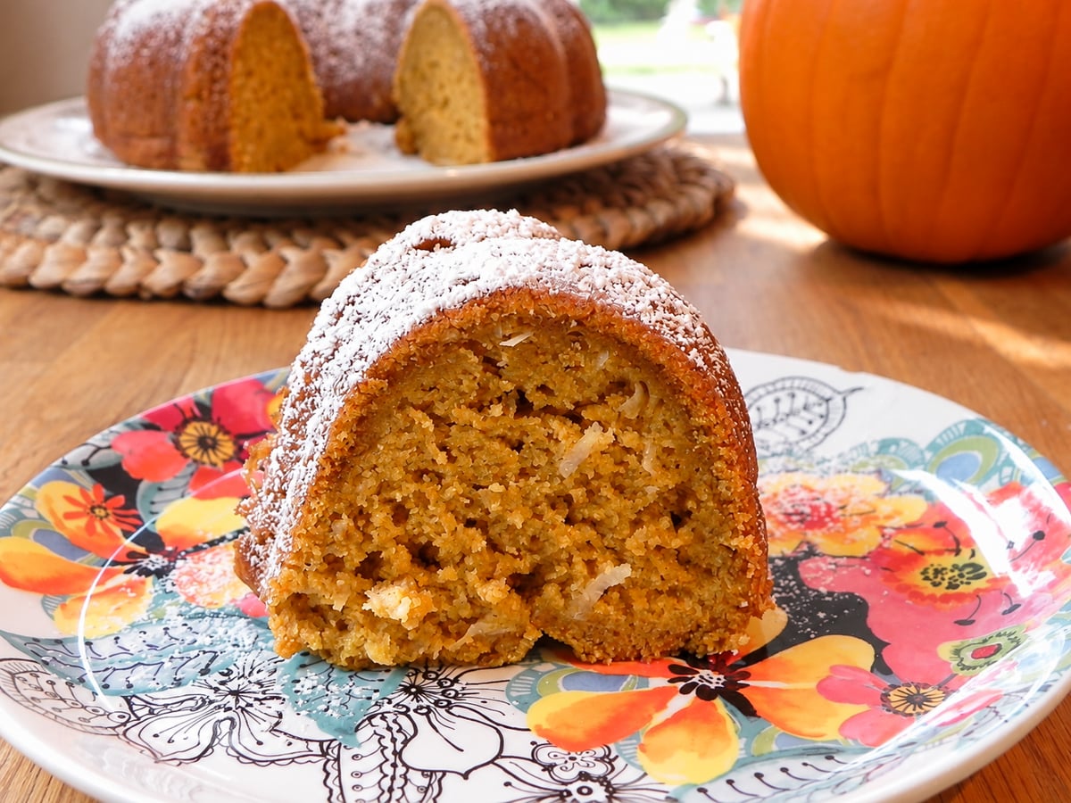 A slice of pumpkin cake dusted with powdered sugar on a colorful floral plate, with a whole pumpkin and the rest of the bundt cake in the background.