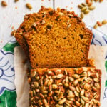 Sliced Sourdough Discard Pumpkin Bread topped with pumpkin seeds on a decorative plate, with extra seeds scattered on a white wooden surface.
