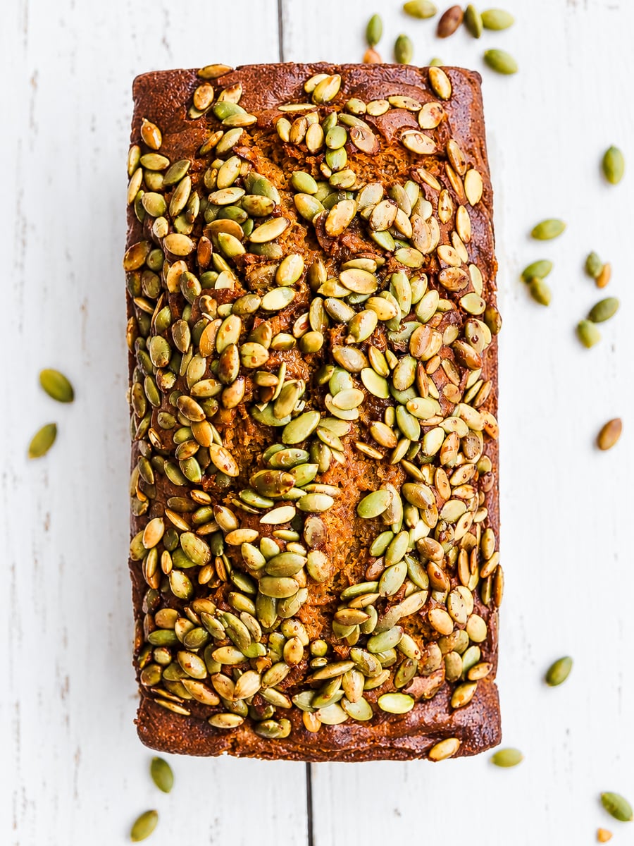 A loaf of Sourdough Discard Pumpkin Bread topped with pumpkin seeds sits on a white wooden surface, with some seeds scattered around.