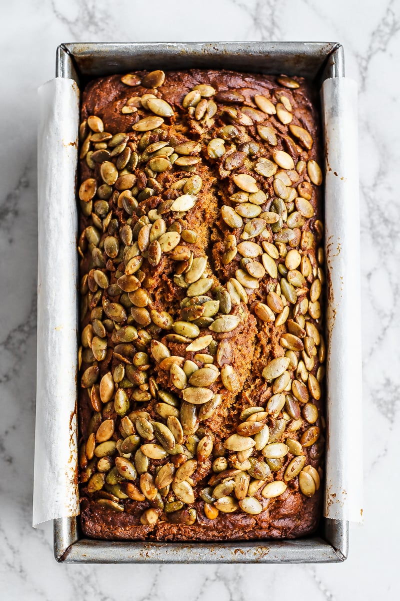 A loaf of Sourdough Discard Pumpkin Bread topped with pumpkin seeds sits in a parchment-lined metal loaf pan on a marble surface.