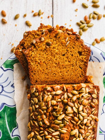 Sliced Sourdough Discard Pumpkin Bread topped with pumpkin seeds on a decorative plate, with extra seeds scattered on a white wooden surface.