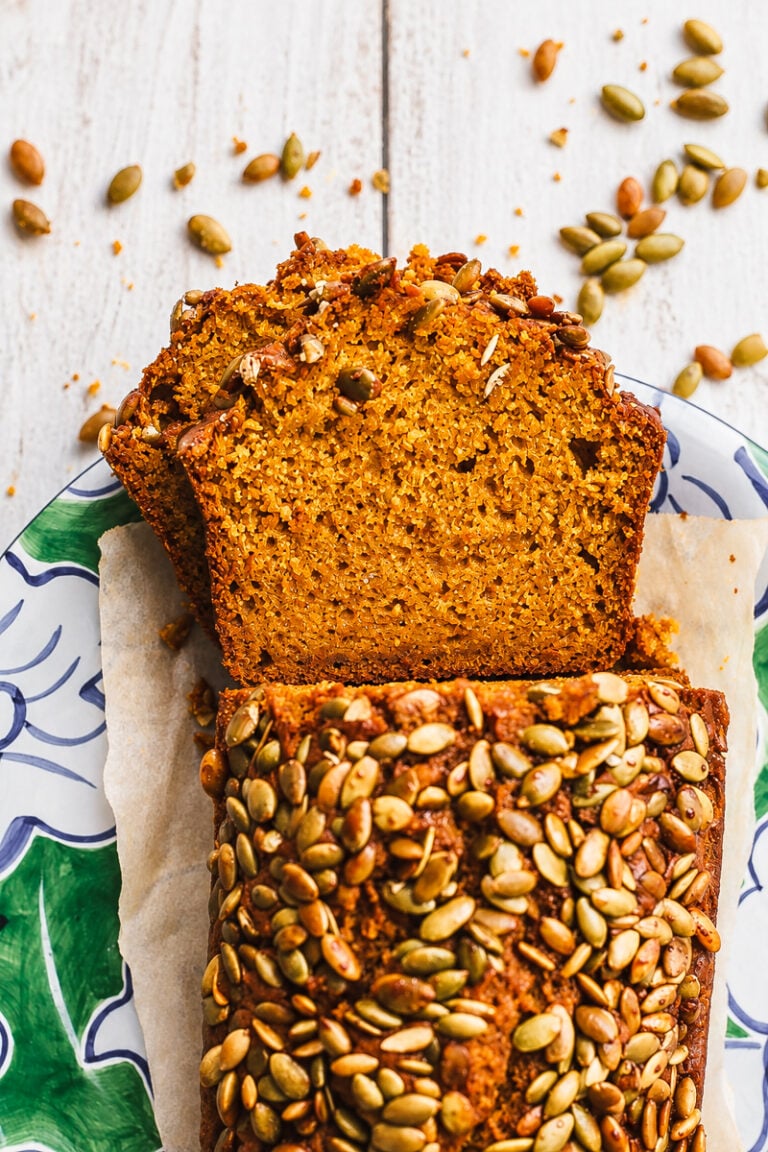 Sliced Sourdough Discard Pumpkin Bread topped with pumpkin seeds on a decorative plate, with extra seeds scattered on a white wooden surface.