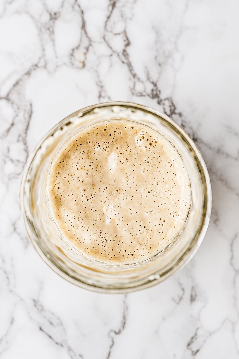 A bubbly sourdough starter in a glass jar sits on a white marble surface, ready to inspire your next bake&mdash;like a delicious Sourdough Discard Pumpkin Bread.