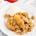 A plate with Easy Apple Crisp with Oats Topping, served warm and finished with a scoop of creamy vanilla ice cream. Two fresh apples and cinnamon sticks in the background complete this inviting dessert scene.