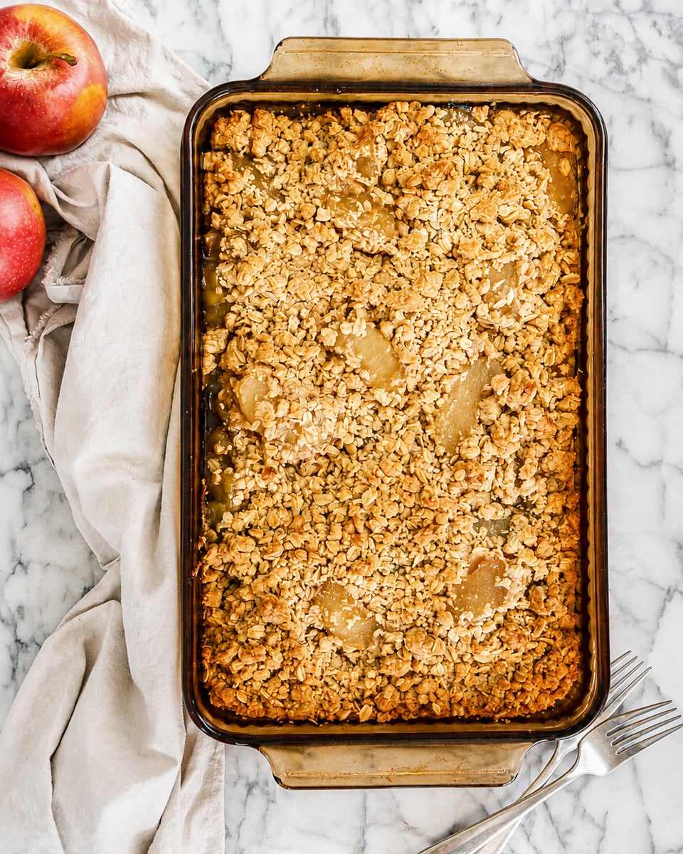 A rectangular pan of Easy Apple Crisp with Oats Topping sits on a marble surface beside a beige cloth, two forks, and two red apples. The crisp features a golden, oat-topped crust.