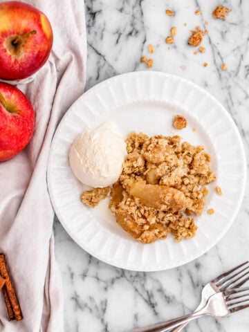 A white plate with Easy Apple Crisp with Oats Topping and a scoop of vanilla ice cream, next to two red apples, cinnamon sticks, and forks on a marble surface.