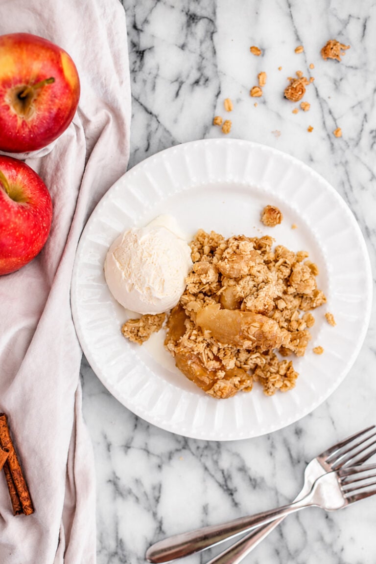 A white plate with Easy Apple Crisp with Oats Topping and a scoop of vanilla ice cream, next to two red apples, cinnamon sticks, and forks on a marble surface.