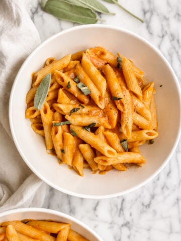 A white bowl filled with creamy penne pasta in a rich orange pumpkin pasta sauce, garnished with fresh sage leaves, sits on a marble surface next to a cloth napkin and extra sage leaves.