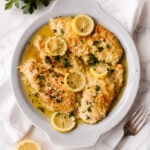 A white plate holds lemon chicken-breaded chicken breasts in a zesty lemon butter sauce, garnished with parsley and lemon slices. Fresh parsley, lemon halves, and a fork are nearby on a white marble surface.