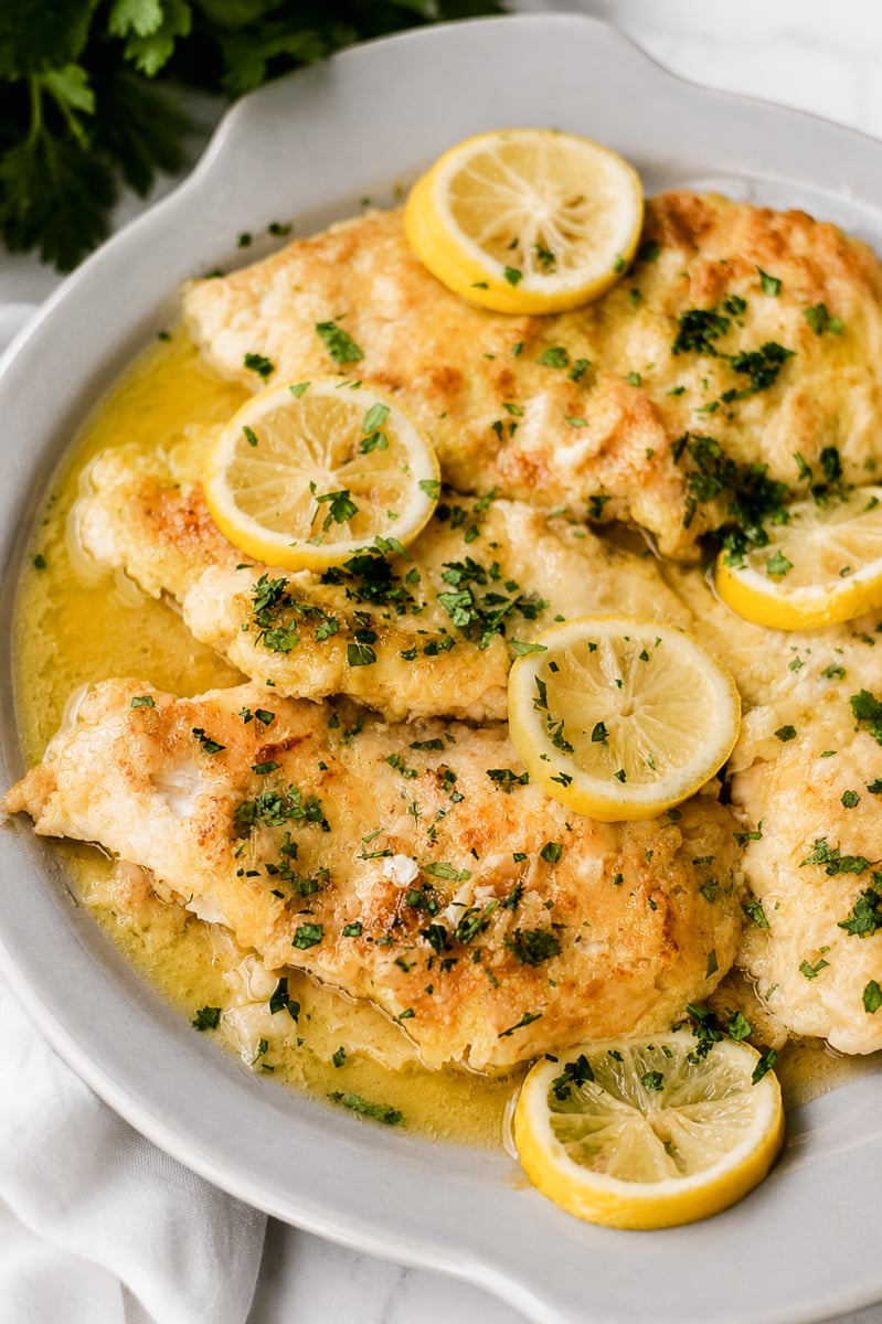 A plate of breaded lemon chicken breasts garnished with chopped parsley and lemon slices, served in a rich lemon butter sauce.