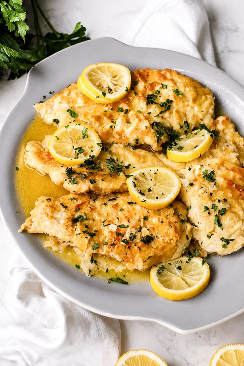 A plate of breaded, baked fish fillets garnished with lemon slices and chopped parsley, reminiscent of lemon chicken, served on a light gray dish with a white cloth and fresh parsley in the background.