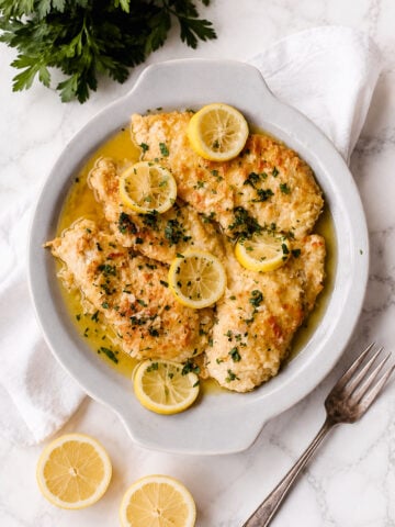 A white plate holds lemon chicken-breaded chicken breasts in a zesty lemon butter sauce, garnished with parsley and lemon slices. Fresh parsley, lemon halves, and a fork are nearby on a white marble surface.