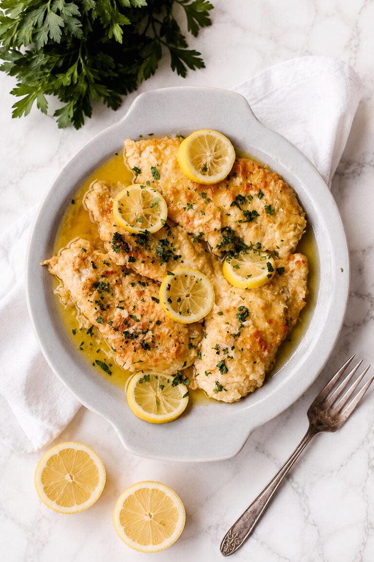 A white plate holds lemon chicken—breaded chicken breasts in a zesty lemon butter sauce, garnished with parsley and lemon slices. Fresh parsley, lemon halves, and a fork are nearby on a white marble surface.