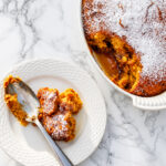 A white dish of golden, moist Pumpkin Pudding Cake dusted with powdered sugar, with a portion served on a white plate and a spoon, on a marble surface.