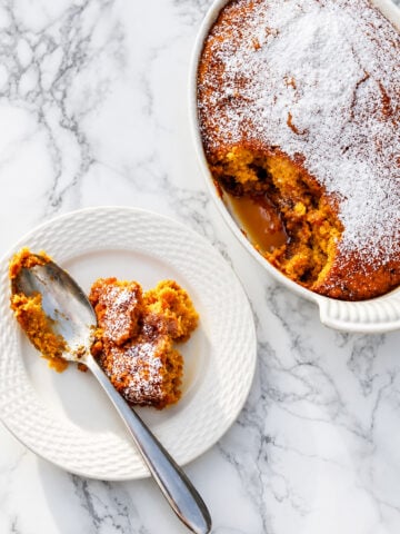 A white dish of golden, moist Pumpkin Pudding Cake dusted with powdered sugar, with a portion served on a white plate and a spoon, on a marble surface.