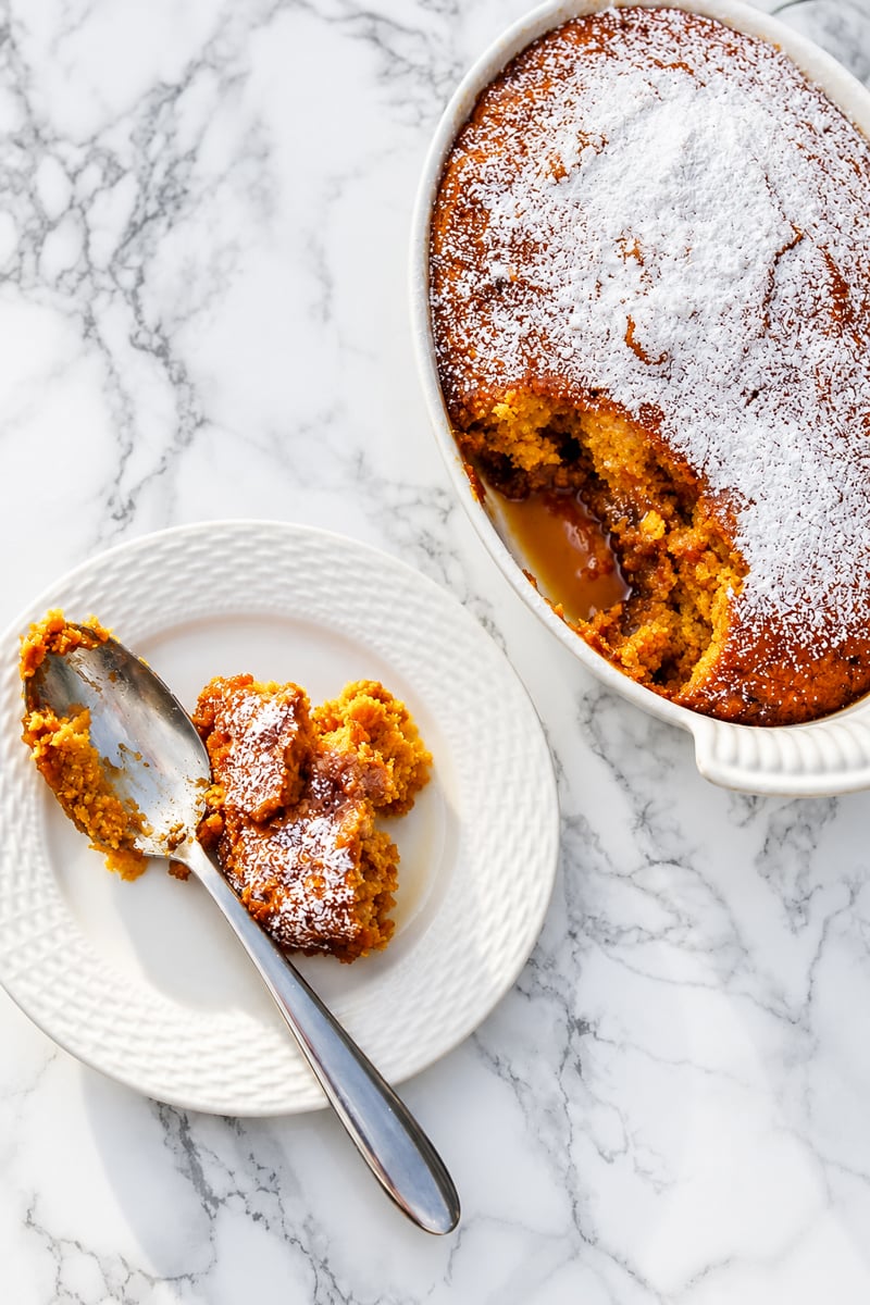 A white dish of golden, moist Pumpkin Pudding Cake dusted with powdered sugar, with a portion served on a white plate and a spoon, on a marble surface.