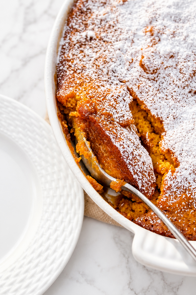 A spoon scoops a portion of golden Pumpkin Pudding Cake, dusted with powdered sugar, from a white dish beside empty plates on a marble surface.