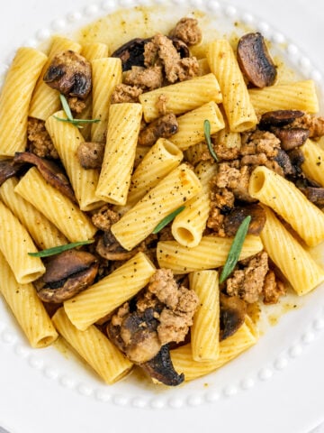 Rigatoni pasta with ground turkey, sautéed mushrooms, fresh herbs, and black pepper—this comforting dish is elevated with a hint of pumpkin, making for a delicious ground turkey pasta with pumpkin. Served on a white plate with a decorative edge.