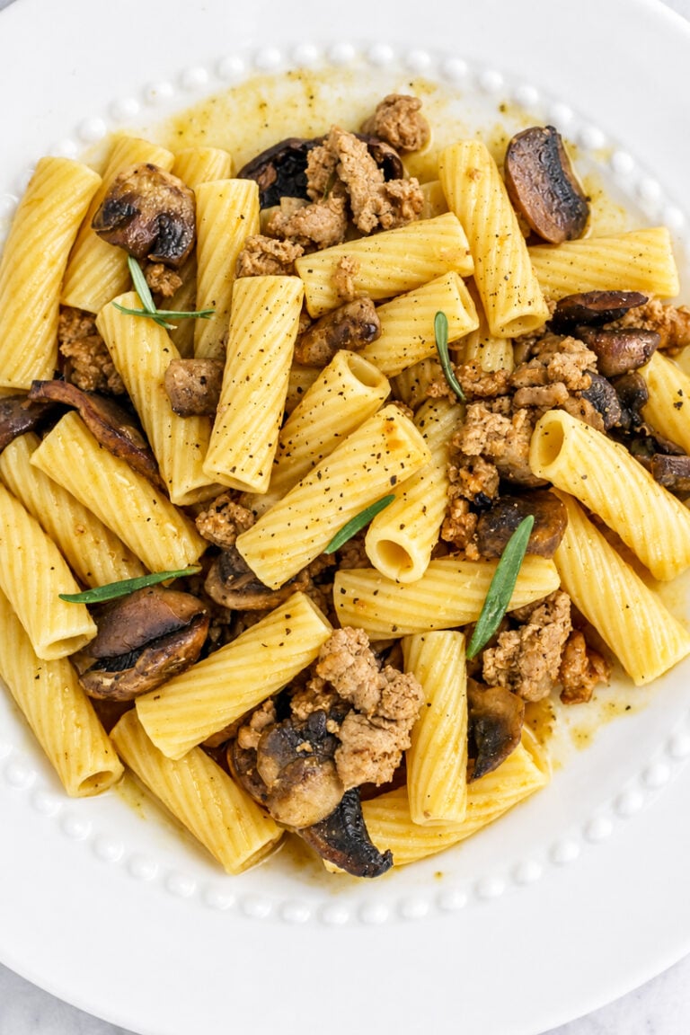 Rigatoni pasta with ground turkey, sautéed mushrooms, fresh herbs, and black pepper—this comforting dish is elevated with a hint of pumpkin, making for a delicious ground turkey pasta with pumpkin. Served on a white plate with a decorative edge.