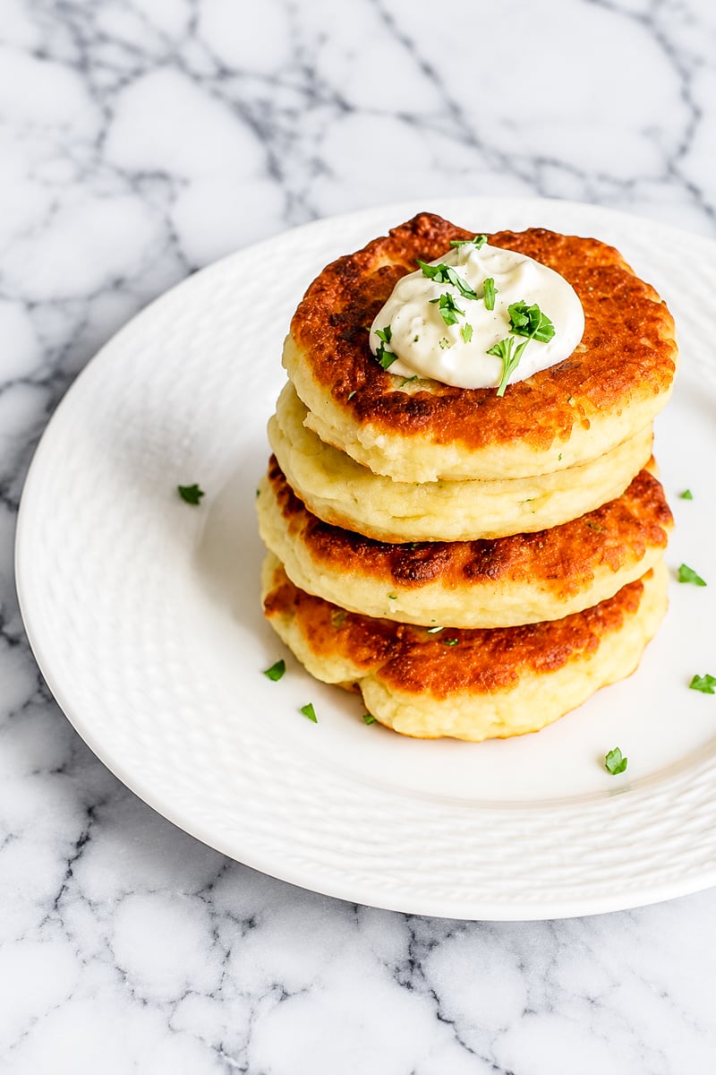 A stack of three golden-brown potato pancakes topped with sour cream and chopped herbs sits on a white plate, set atop a marble surface.