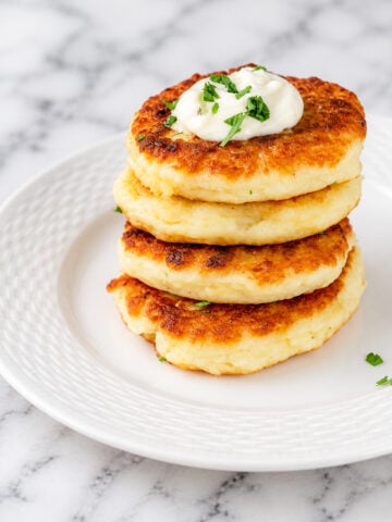 Three golden brown potato pancakes stacked on a white plate, topped with a dollop of sour cream and garnished with chopped fresh herbs. The classic potato pancakes rest on a marble surface.