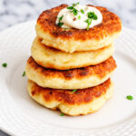 Four golden-brown potato pancakes stacked on a white plate, topped with a dollop of sour cream and chopped herbs, are displayed on a marble surface.