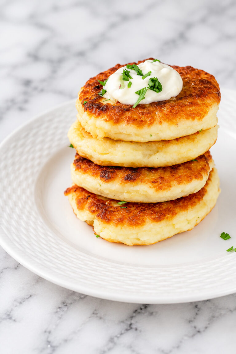 Three golden brown potato pancakes stacked on a white plate, topped with a dollop of sour cream and garnished with chopped fresh herbs. The classic potato pancakes rest on a marble surface.