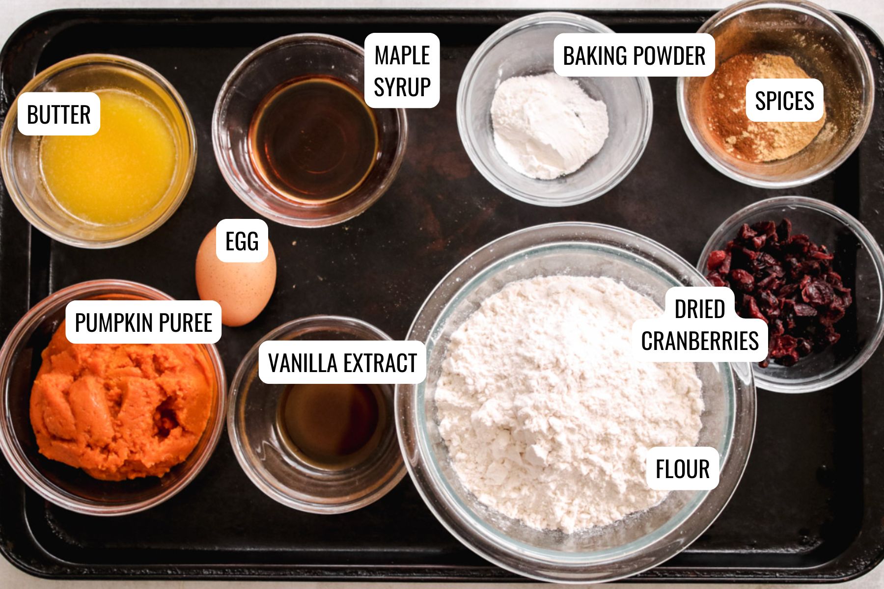 A baking tray holds bowls with labeled ingredients for Pumpkin Cranberry Scones: melted butter, pumpkin puree, an egg, vanilla extract, maple syrup, flour, baking powder, spices, and dried cranberries.