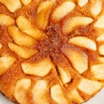 A golden-brown Apple Upside-Down Cake topped with neatly arranged glazed apple slices sits on a white plate.