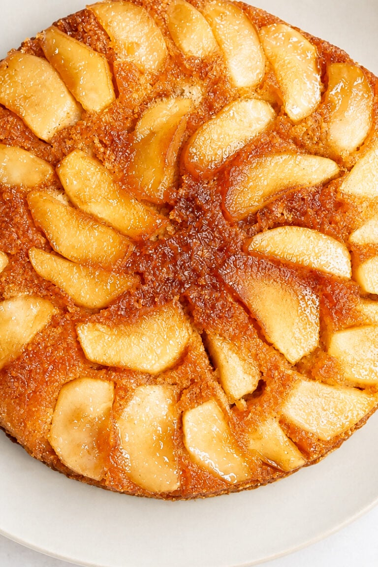 A golden-brown Apple Upside-Down Cake topped with neatly arranged glazed apple slices sits on a white plate.