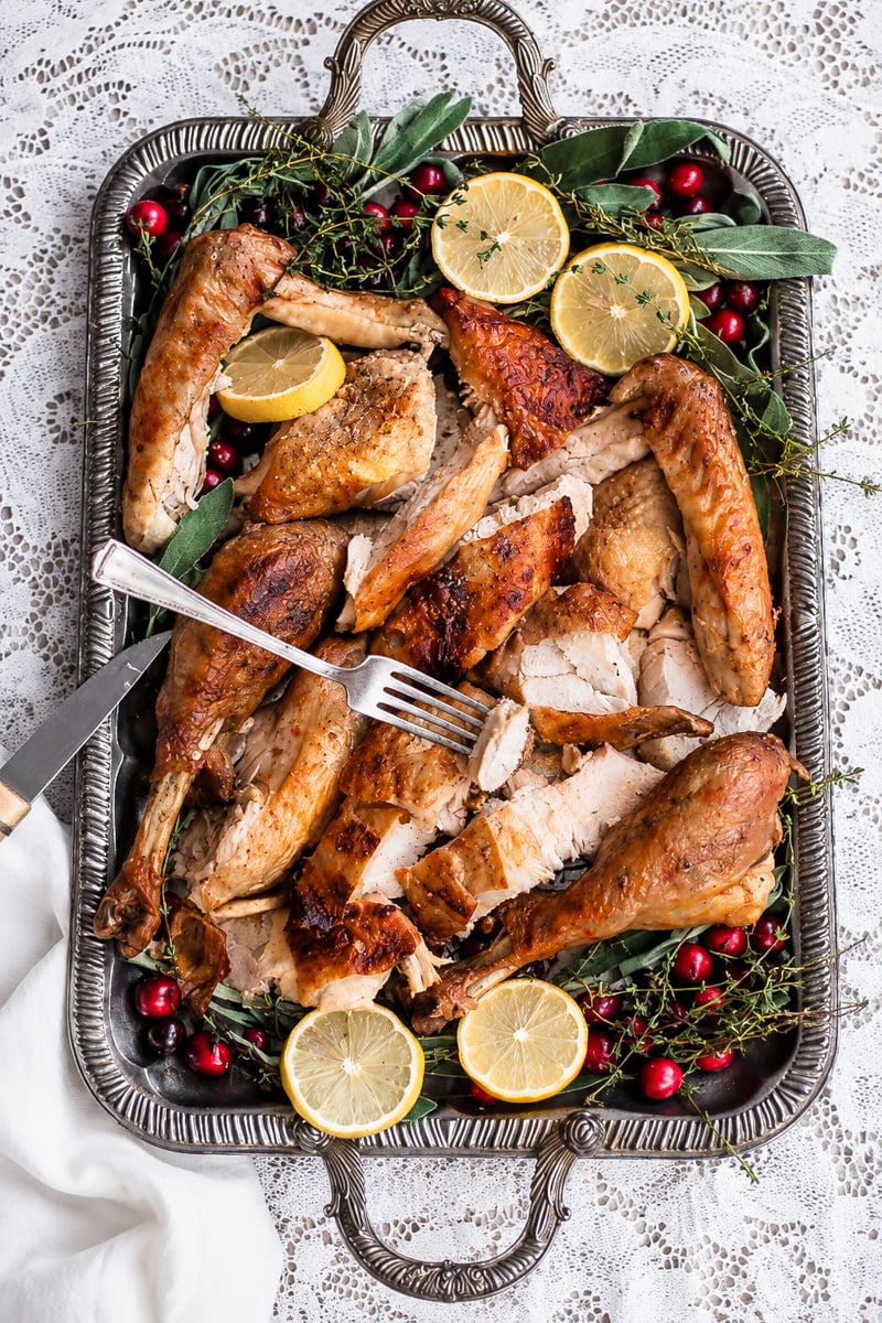 A silver tray with perfectly carved roast turkey, lemon slices, fresh sage, thyme, and cranberries rests on a lace tablecloth. A fork and carving knife are elegantly arranged on the tray.