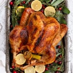 A beautifully prepared roast turkey sits on a silver tray, garnished with lemon slices, fresh herbs, and cranberries, perfectly displayed on a white lace tablecloth.