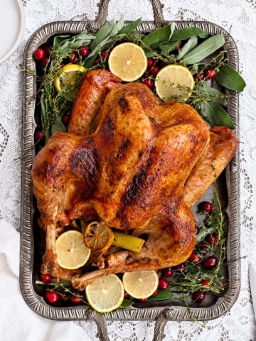 A beautifully prepared roast turkey sits on a silver tray, garnished with lemon slices, fresh herbs, and cranberries, perfectly displayed on a white lace tablecloth.