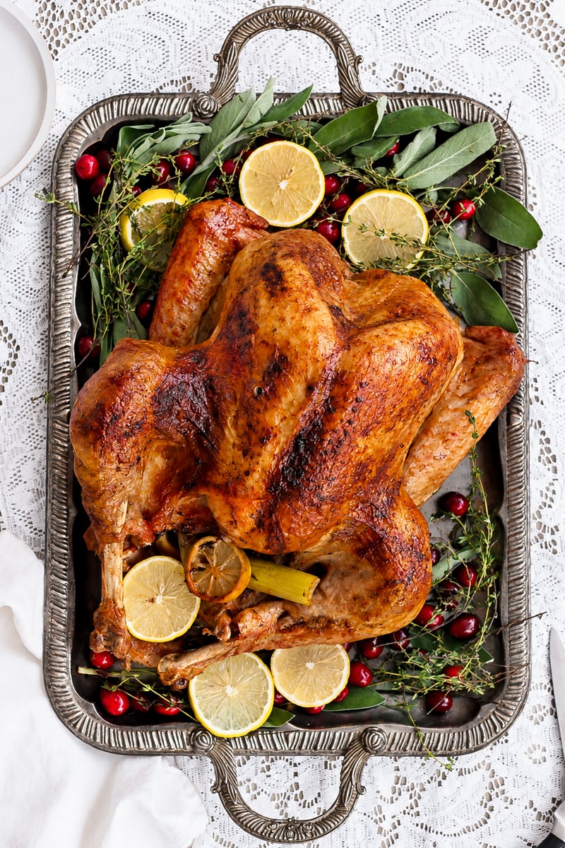 A beautifully prepared roast turkey sits on a silver tray, garnished with lemon slices, fresh herbs, and cranberries, perfectly displayed on a white lace tablecloth.