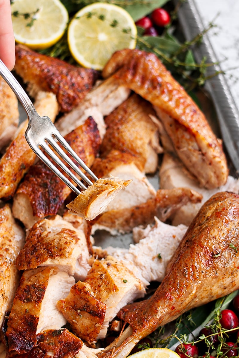 A fork holds a piece of roast turkey above a platter with sliced turkey breast, drumsticks, lemon slices, cranberries, and herbs.
