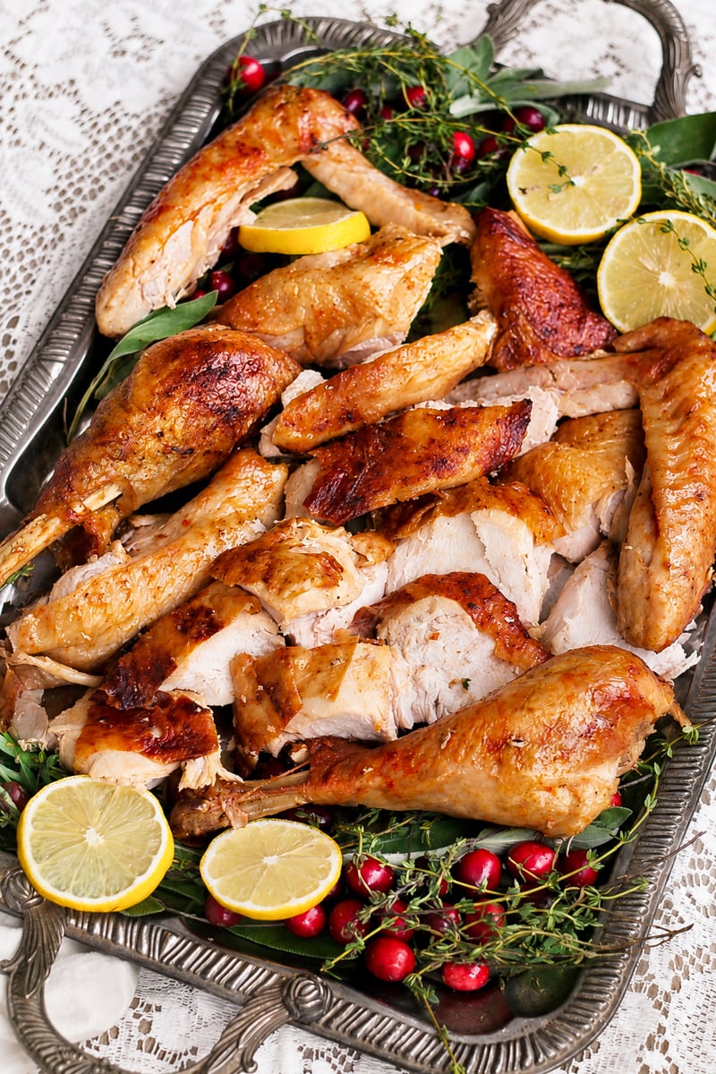 Sliced roast turkey arranged on a tray with fresh lemon slices, cranberries, herbs, and leafy greens, set on a lace tablecloth for an elegant presentation of classic roast turkey.