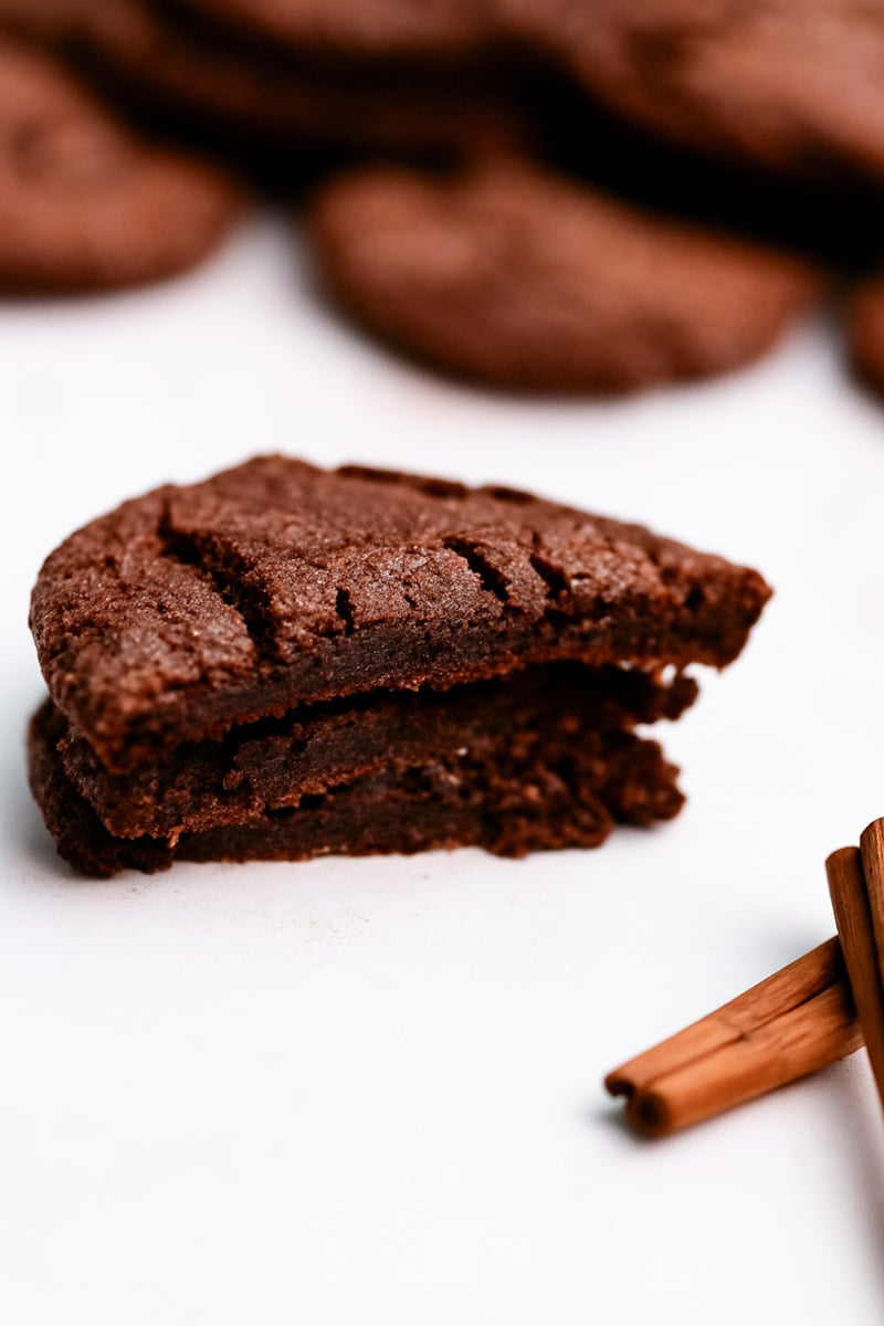 A close-up of a Vegan Mexican Chocolate Snickerdoodle cookie broken in half, showing its soft, fudgy interior. In the foreground are two cinnamon sticks, with more cookies blurred in the background.