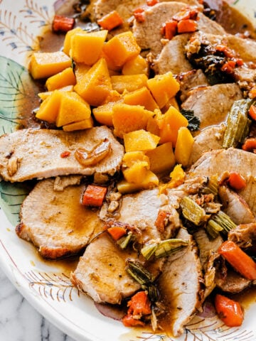Sliced Roast Pork Tenderloin with Butternut Squash, accompanied by carrots, celery, and chunks of cooked yellow squash on a decorative plate, all topped with savory brown sauce.