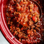 A ladle scoops hearty chili with ground beef, tomatoes, and vegetables from a red slow cooker, much like the comforting process of preparing Slow Cooker Turkey Bolognese Sauce.