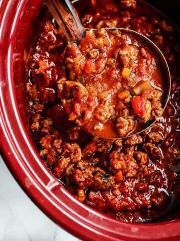 A ladle scoops hearty chili with ground beef, tomatoes, and vegetables from a red slow cooker, much like the comforting process of preparing Slow Cooker Turkey Bolognese Sauce.