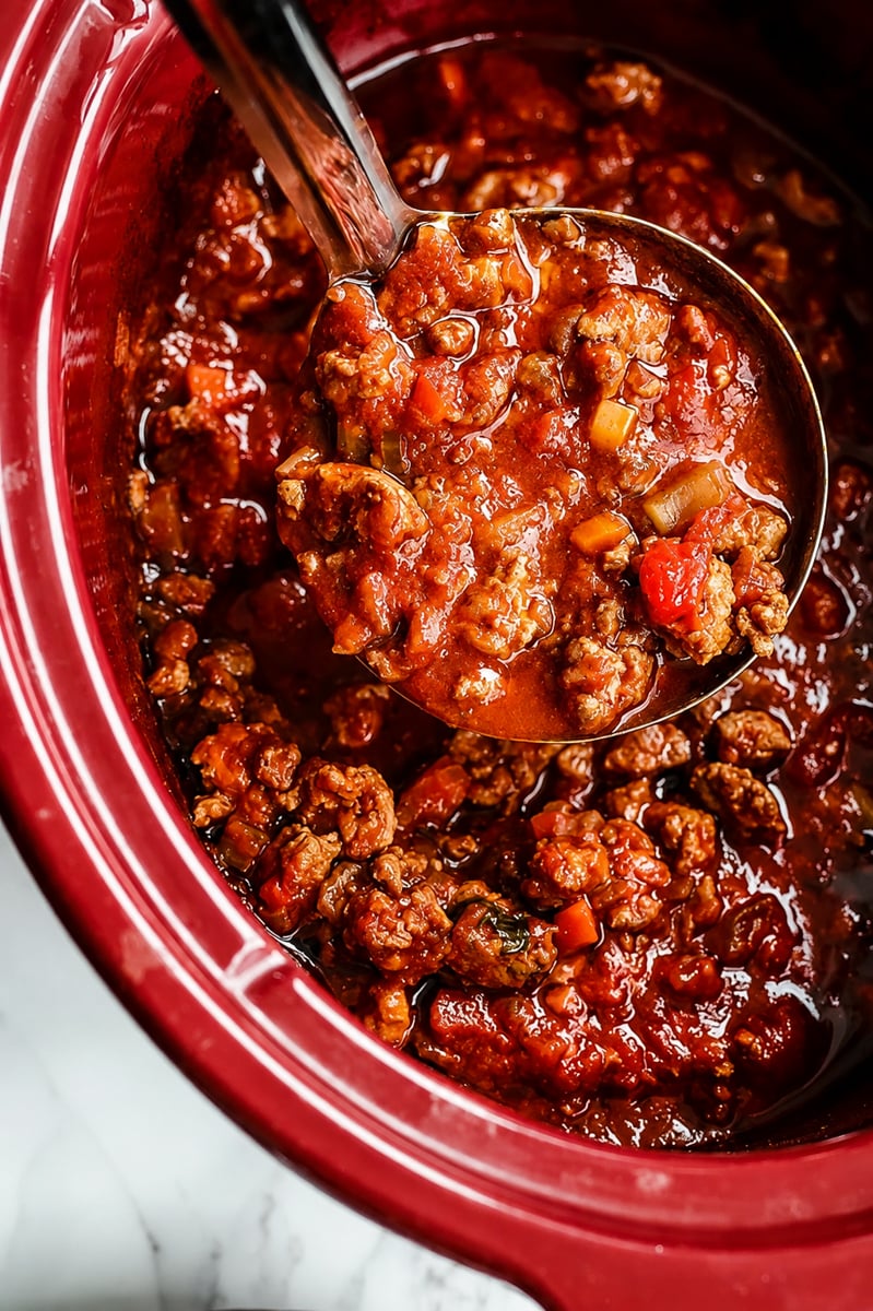 A ladle scoops hearty chili with ground beef, tomatoes, and vegetables from a red slow cooker, much like the comforting process of preparing Slow Cooker Turkey Bolognese Sauce.