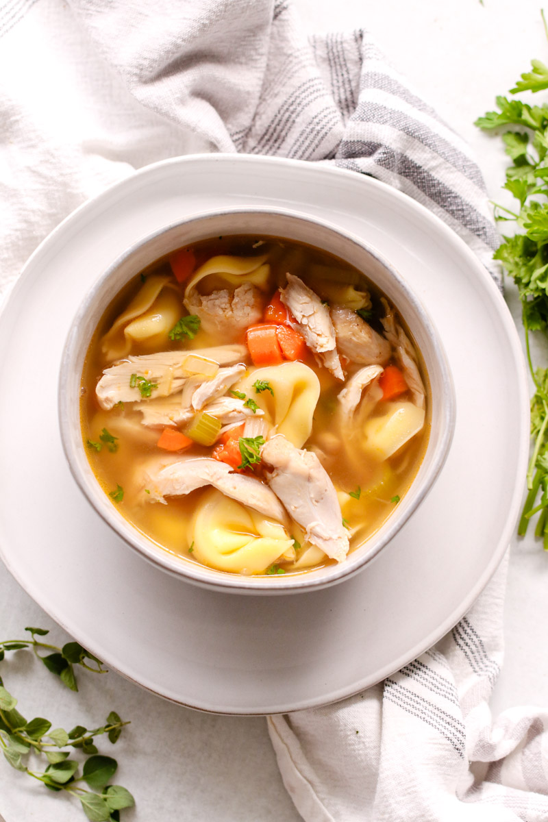 overhead image of chicken tortellini soup in a bowl with herbs in the background. 