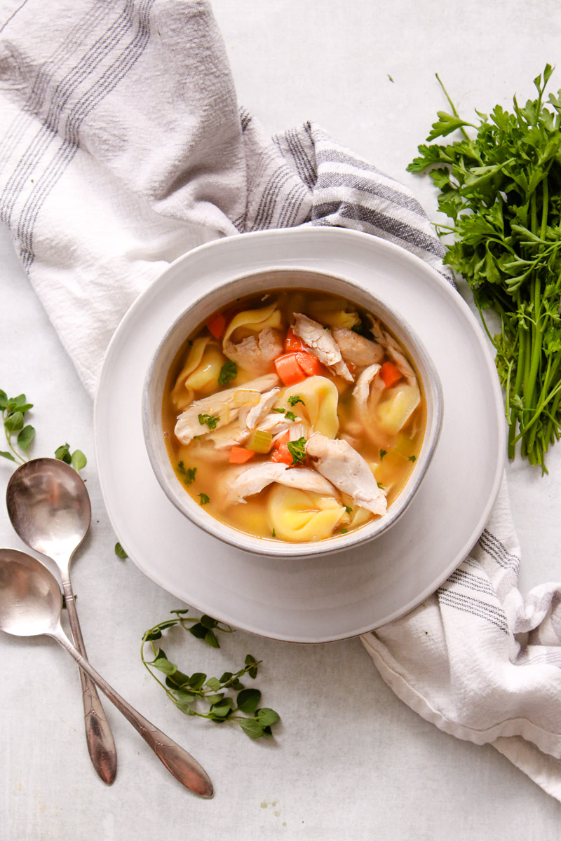 overhead image of chicken tortellini soup in a bowl with herbs in background.