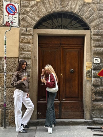 Two women stand in front of a large wooden door set in a stone building, holding drinks and conversing-perhaps sharing tips on seeing Florence in One Day. Street signs and a house number 26 plaque are visible on the wall beside them.