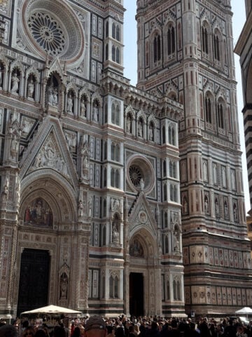 The ornate facade of Florence Cathedral and its bell tower, featuring intricate marble designs and sculptures, greets a lively crowd-an essential stop for anyone exploring Florence in One Day under sunny skies.