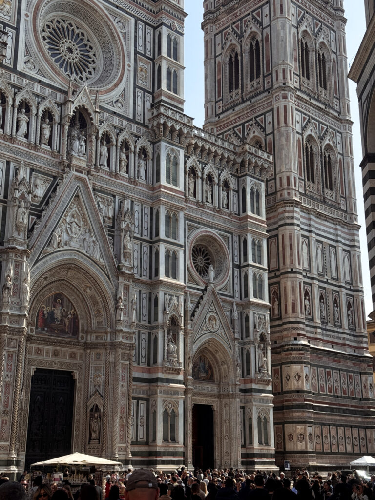 The ornate facade of Florence Cathedral and its bell tower, featuring intricate marble designs and sculptures, greets a lively crowd—an essential stop for anyone exploring Florence in One Day under sunny skies.