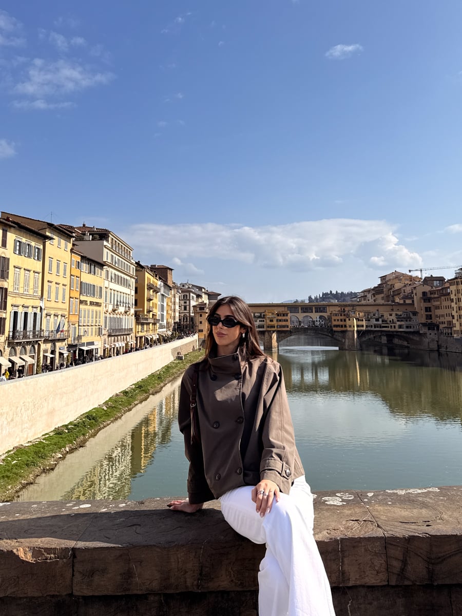 A woman wearing sunglasses and a brown jacket sits on a stone railing by a river with colorful buildings and the Ponte Vecchio bridge in Florence, Italy-perfect inspiration for exploring Florence in One Day under blue skies with scattered clouds.